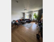 Helles Wohnzimmer mit breitem Holzdielenboden, schwarzem Ecksofa und Sessel um einen niedrigen Couchtisch auf einem Teppich. Zwei große Fenster mit Vorhängen, ein Heizkörper darunter, mehrere Zimmerpflanzen sowie eine Wand mit gerahmten Fotos prägen den Raum.