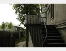 Außenansicht eines alten Gebäudes mit einer schmalen Metalltreppe, die zu einer überdachten Eingangstür führt. Im Hintergrund ist ein weißes Haus mit Bäumen und eine von Hecken und Zaun gesäumte Grünfläche erkennbar.