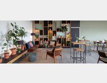 Ein gemütlicher, modern eingerichteter Innenraum mit einer Mischung aus Lounge- und Essbereich, bepflanzten Regalen an einer Wand, Holzmöbeln, verschiedenen Zimmerpflanzen und einem Teppich auf einem gefliesten Boden. Links stehen kleine Sofas und Sessel, rechts hohe Tische und Stühle.