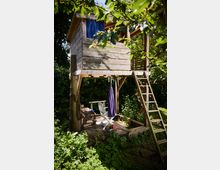 Holz-Baumhaus auf Stelzen in einem dicht bewachsenen Garten, mit kleiner Veranda, seitlicher Leiter und blauem Vorhang am Fenster. Darunter befindet sich eine Holzplattform mit Sitzmöbeln und Sonnenschirm, umgeben von Bäumen und Sträuchern.