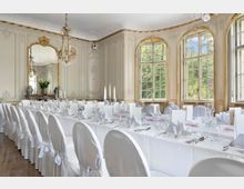 Festlich dekorierter Speisesaal in einem historischen, barocken Gebäude mit reich verzierten Stuckwänden, goldenen Verzierungen und großen, bodentiefen Fenstern. Ein langer Tisch ist mit weißen Tischdecken, gefalteten Stoffservietten, Kerzenleuchtern und eleganten Gläsern gedeckt.