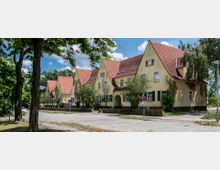 Reihenhäuser mit gelben Fassaden und roten Satteldächern entlang einer Kopfsteinpflasterstraße, umgeben von Bäumen und kleinen Gärten. Im Vordergrund stehen Straßenschilder und Laternen.