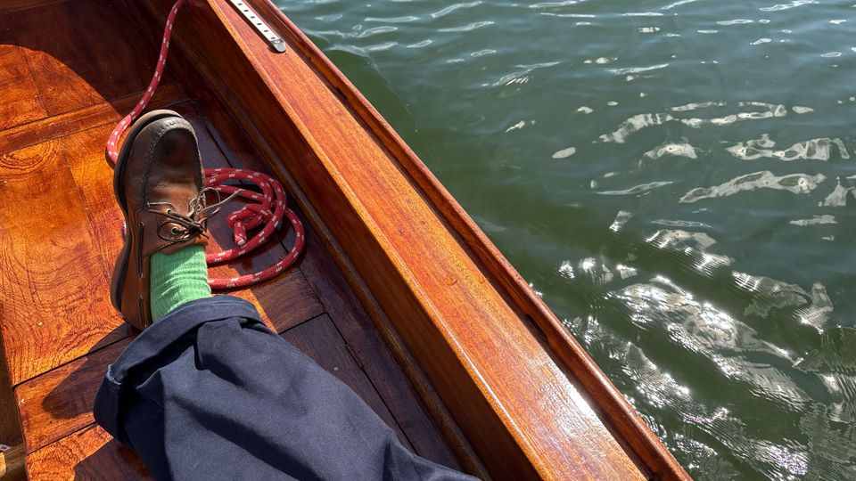 Blick in ein hölzernes Boot mit glänzendem Deck und Bordwand; ein rotes Seil liegt im Boot. Daneben ist die Wasseroberfläche mit leichten Wellen und Sonnenreflexen zu sehen.