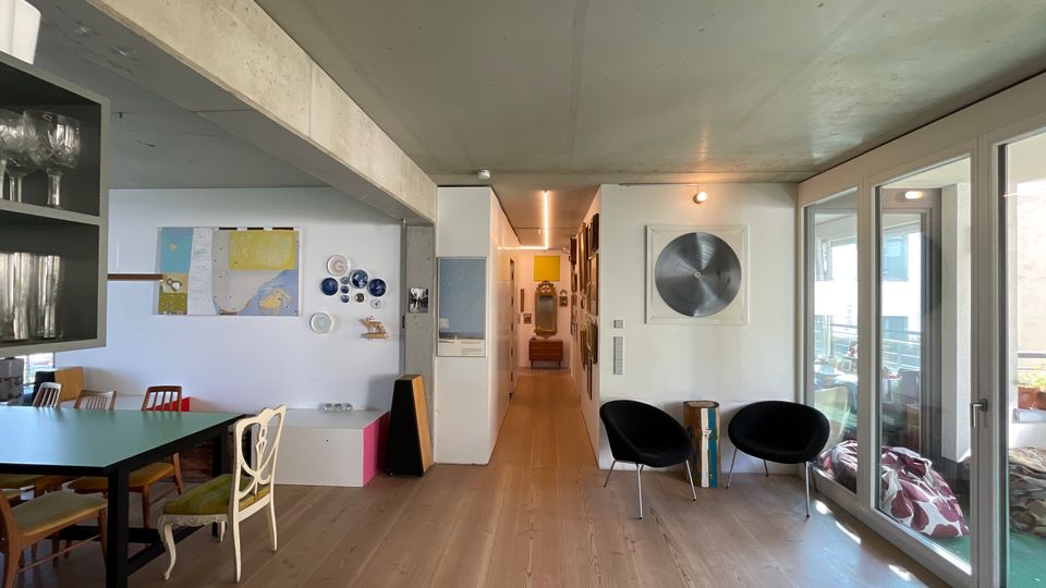 Wohnbereich in einem modernen Innenraum mit heller Holzfußboden, einer langen Tafel mit unterschiedlichen Stühlen und einer Galerie aus Dekorplatten an der Wand. Auf der rechten Seite befinden sich große Glasfenster, die zu einem Balkon führen.