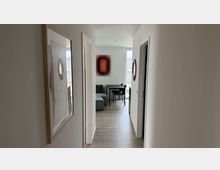 Schmaler, heller Flur einer modernen Wohnung mit weißen Wänden, Holzfußboden und mehreren Türöffnungen, an den Wänden hängen gerahmte Kunstwerke. Am Ende öffnet sich der Flur zu einem Wohn- und Essbereich mit grauem Sofa, kleinem Esstisch und großen Fenstern mit Blick nach draußen.