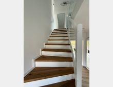 Treppe in einem modernen Innenraum mit Holzstufen und weißen Seitenwangen, rechts ein Geländer mit Glasverkleidung. Im oberen Bereich führt die Treppe zu einer weißen Tür, begleitet von minimalistischem Dekor und neutralem Licht.