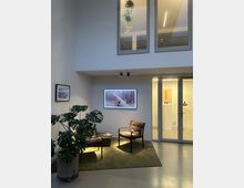 Moderner Innenraum mit einer kleinen Sitzecke, bestehend aus zwei gepolsterten Stühlen auf einem grünen Teppich und einer Zimmerpflanze in einem Topf. Im Hintergrund hängen ein großes beleuchtetes Foto und ein kleineres Bild an der Wand; rechts befinden sich Glaswände und -türen, die in angrenzende Räume führen.