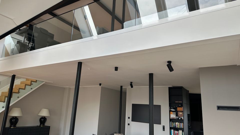 Moderner, offener Wohnraum mit hoher Decke, sichtbaren schwarzen Stahlträgern und einer Galerie mit Glasgeländer. Unten sind ein großes graues Sofa, eine Treppe zur Galerie, dezente Einbaubeleuchtung sowie ein Regalbereich neben einem Durchgang zu sehen.