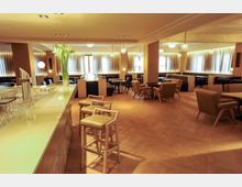 Modern eingerichteter Loungebereich mit hellem Holzboden, gepolsterten Sesseln und Tischen, einer Theke mit Barhockern im Vordergrund sowie großen Fenstern mit Vorhängen, die warmes, weiches Licht hereinlassen. Deckenspots und Hängeleuchten sorgen für zusätzliche Beleuchtung.