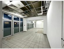 Leerer Raum mit weißen Wänden, gefliesten Böden und großen Glasfenstern sowie -türen an einer Seite. Die Decke zeigt freiliegende Kabel und Lüftungsrohre in einem offenen Rasterdesign.