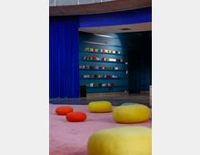 Innenansicht eines modernen Raums mit bunten Sitzkissen auf einem rosa Teppichboden. Im Hintergrund befindet sich eine dunkelblaue Wand mit horizontalen Regalböden, die mit kleinen bunten Büchern oder Objekten bestückt sind, abgetrennt durch einen großen blauen Vorhang.