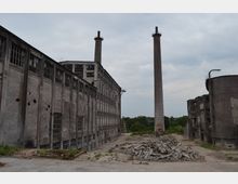 Verlassenes Industriegebiet mit alten, grauen Fabrikgebäuden, zwei hohen Schornsteinen und Trümmerhaufen auf einem asphaltierten Platz. Im Hintergrund sind Bäume und bewölkter Himmel sichtbar.