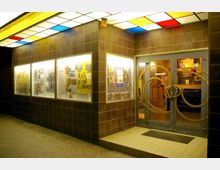 Eingang eines kleinen Kinos mit dunkel gefliester Fassade, hell beleuchteten Schaukästen mit Filmplakaten und Glastüren mit auffälligen Metallornamenten. Über dem Eingang befindet sich eine Deckenbeleuchtung mit bunten, quadratischen Glasfeldern.