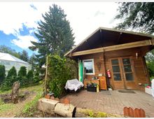 Ein kleines Holzhaus mit Terrasse, umgeben von grünem Garten mit Bäumen und Sträuchern. Im Hintergrund ist ein Gewächshaus und eine große Tanne sichtbar, der Boden ist mit gepflasterten Steinen bedeckt.