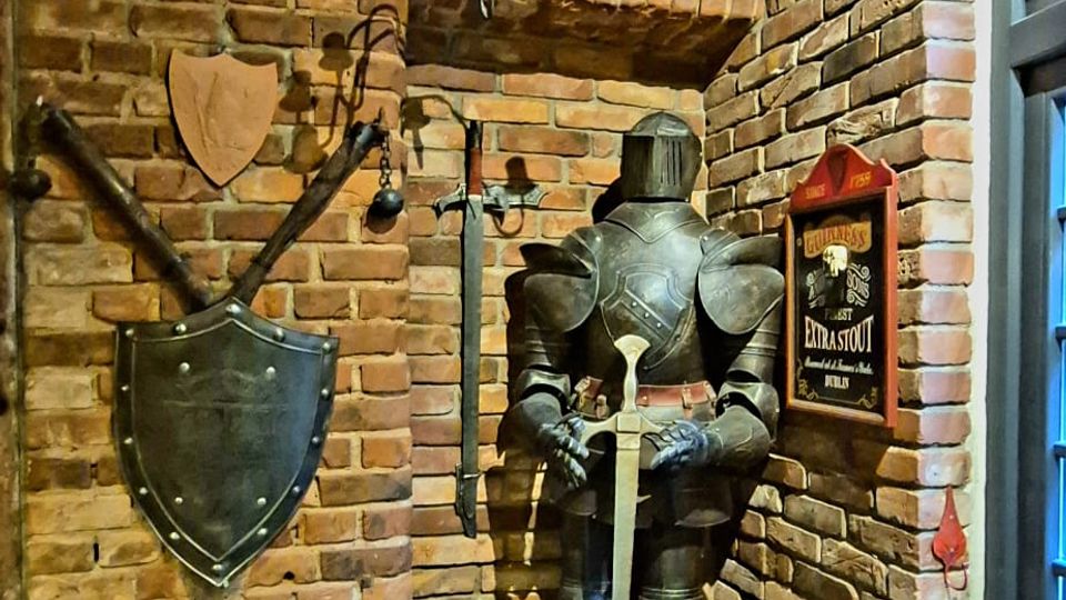 Rustikales Innenraum-Eck mit unverputzten Backsteinwänden, an denen ein Schild und gekreuzte Waffen dekorativ angebracht sind. In einer Nische steht eine Ritterrüstung mit Schwert unter einem schmiedeeisernen Kronleuchter, daneben ein Fenster und Holzmöbel im Vordergrund.