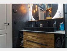 Modernes Badezimmer mit Waschtisch aus Holz und einem schwarzen Waschbecken. Im Hintergrund sind dunkelgraue Wandfliesen, ein beleuchteter runder Spiegel und hängende Glühbirnen zu sehen.