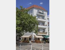 Mehrstöckiges Wohngebäude mit weißer Fassade und rotem Ziegeldach, Balkonen mit Pflanzen und Sonnenschirmen. Im Erdgeschoss befindet sich ein Café mit Markisen, Holzbänken und Sitzbereichen im Freien, daneben Bäume und eine gepflasterte Straße.