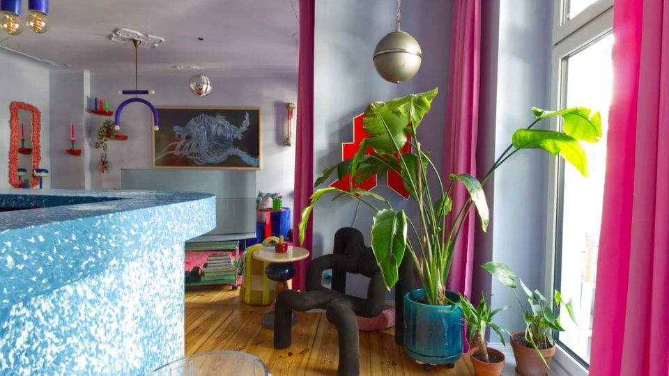 Bright, colorful living room with large windows framed by pink curtains, a wooden floor, and several potted plants including a tall leafy plant in a blue pot. The space features playful modern decor such as sculptural black seating, a small round side table, hanging globe lights, and wall art in pastel tones.
