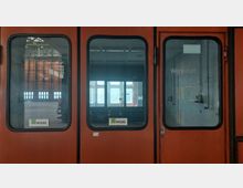 Orangefarbene Doppeltür mit Fenstern, die in eine Werkstatt führt. Auf den Scheiben befinden sich Beschriftungen, darunter das Wort „Werkstatt“ und ein „WISAG“-Logo; im Hintergrund ist ein großer, heller Raum mit Fenstern und industriellen Strukturen sichtbar.