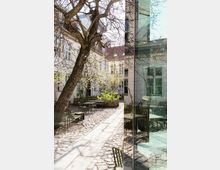 Innenhof mit Kopfsteinpflaster und Sitzgelegenheiten, umgeben von historischen Gebäuden mit weißen Fassaden; ein großer, blattloser Baum steht zentral im Hof, während eine spiegelnde Glasfläche rechts im Bild moderne Architektur zeigt.