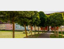 Mehrfarbige Reihenhäuser mit pastellfarbenen Fassaden (gelb, rosa, hellblau) stehen hinter einer Grünfläche mit jungen Bäumen. Ein schmaler gepflasterter Weg führt durch die Bäume zu einem Torbogen zwischen den Gebäuden.