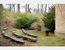 Freiluft-Andachtsplatz mit halbkreisförmig angeordneten Holzsitzbänken, einem Holztisch und einem Baumstumpf mit eingeschnitztem Kreuzmotiv, umgeben von Bäumen, Sträuchern und einer Backsteinmauer an der rechten Bildseite.