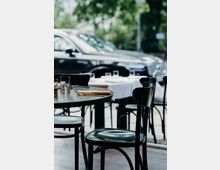 Ein Café mit schwarzen Holzmöbeln, gedeckten Tischen, Gläsern und Servietten; im Hintergrund ist ein geparktes Auto und verschwommene grüne Bäume zu sehen.