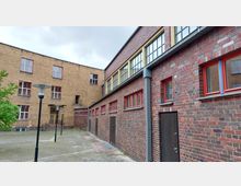 Backsteinfassade eines Gebäudekomplexes mit roten Fensterrahmen und großen, teilweise vergitterten Fenstern. Im Vordergrund eine gepflasterte Fläche mit Laternen; links im Bild ist ein kleiner Baum sichtbar.