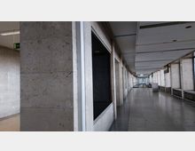 Ein leerer, länglicher Flur in einem Gebäude mit hellen Natursteinwänden, großen Fenstern auf der rechten Seite und einer gefliesten Bodenfläche. Die Decke zeigt eingelassene Beleuchtungselemente, während am Ende des Flurs Glastüren und eine große Uhr sichtbar sind.