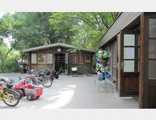 Eine Außenaufnahme eines Spiel- und Fahrradverleihs mit zwei Holzhütten, Kinderfahrzeugen und Fahrrädern. Im Vordergrund stehen bunte Fahrradhelme auf einem Ständer, umgeben von Bäumen und gepflastertem Boden.