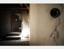 Blick in einen alten, verlassenen Innenraum mit abgenutzten Wänden, einer Holztreppe und einer offenen Holztür, durch die Licht einfällt. Sichtbare Holzbalken und Risse in der Wand betonen den verfallenen Zustand.