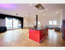 Großzügiger, moderner Raum mit Holzboden, einer roten Kücheninsel in der Mitte und einer Sitzecke mit grauen Sofas. Große Fenster lassen viel Tageslicht herein, ergänzt durch schwarze Vorhänge und Dachfenster auf der rechten Seite.