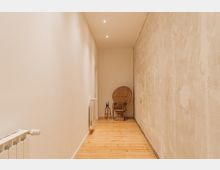 Schmaler Flur mit Holzdielenboden und einer Kombination aus glatten, weißen Wänden auf der linken und verputzten, strukturierten Wänden auf der rechten Seite. Im Hintergrund steht ein runder Rattanstuhl neben einem kleinen Tisch mit einer Vase.