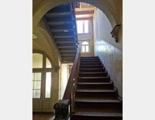 Treppenhaus in einem alten Gebäude mit abgenutzten Holzstufen, einem Holzgeländer und einem Fenster mit Rundbogen im oberen Stockwerk, durch das Licht einfällt. Links befindet sich eine doppelflügelige Glastür mit verzierten Rahmen, rechts ist eine farblich verblasste Wand zu sehen.