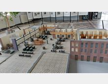 Modell eines historischen Industriegebäudes mit roten Backsteinfassaden, großen Fensterreihen und detaillierter Innenraumaufteilung, einschließlich Maschinen und Ausstattung auf einem Boden mit geometrischem Fliesenmuster. Im Hintergrund befinden sich Ausstellungstafeln und ein ausgestellter Raum mit Fliesenboden.