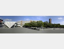 Weitläufiger Platz mit einem modernen, weiß gefalteten Gebäude links, einem langgestreckten Gebäude mit rechteckigen Fenstern im Hintergrund, einem Brunnen mit Wasserfontänen in der Mitte und Bäumen sowie einem Turm im rechten Bildbereich. Das Setting befindet sich in einer städtischen Umgebung mit klarem Himmel.