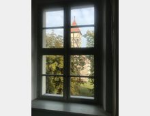Blick durch ein rechteckiges Fenster mit mehreren Sprossen auf einen Kirchturm mit Fachwerkstruktur und rotem Dach, umgeben von Bäumen und einem grasbewachsenen Außenbereich.