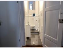 Blick in ein kleines, modernes Badezimmer mit weiß gefliesten Wänden, grauem Fliesenboden, einer wandmontierten Toilette, einem kleinen Waschbecken und einem Heizkörper. Im Hintergrund ist ein Fenster mit Gitter zu sehen.