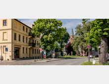 Straßenszene mit einem gelben, historischen Wohn- und Geschäftshaus auf der linken Seite, umgeben von Bäumen. Im Hintergrund ist eine Kirche mit Spitzdach und rotem Ziegelmauerwerk erkennbar, die von weiteren Gebäuden und Grünflächen umgeben ist.