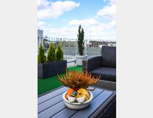 Eine Dachterrasse mit grünem Kunstrasenboden, dekoriert mit Pflanzkübeln und Gartenmöbeln aus grauem Rattan. Im Vordergrund steht auf einem Tisch eine Schale mit Zierkürbissen, Heidekraut und zwei weißen Keramikfiguren in Form von Vögeln.