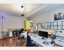 Heller Büroraum mit modernen Möbeln, zwei weißen Schreibtischen und Stühlen, eingerahmt von Regalen mit Büromaterialien. Große Glasfenster an der rechten Wand lassen Tageslicht herein, während der Parkettboden durch bunte Holzstücke akzentuiert ist.