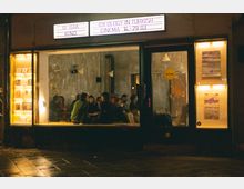 Außenansicht eines kleinen Kinos mit Glasfront, das den Namen „bi'bak“ trägt. Im Inneren sitzen Menschen in einer gemütlichen Atmosphäre vor einer rohen, unverputzten Wand, rechts und links davon beleuchtete Schaukästen mit Büchern und Postern.