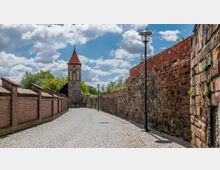 Gepflasterter Weg entlang einer historischen Stadtmauer aus Backstein und Feldsteinen, mit moderner Straßenlaterne und einem roten Kirchturm im Hintergrund, umgeben von Bäumen und einem bewölkten Himmel.