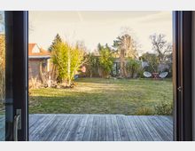 Blick von einem Innenraum durch eine Glastür auf einen Garten mit Holzboden-Terrasse im Vordergrund. Der Garten enthält eine Rasenfläche, einige Bäume und Sträucher, sowie zwei weiße Stühle und einen roten Tisch im Hintergrund.