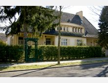 Gelbes Einfamilienhaus im traditionellen Stil mit steilem Dach und mehreren Gauben, umgeben von einer hohen Hecke und einem grünen Metalltor. Im Vordergrund sind Bäume, ein Gehweg und eine Straße zu sehen.