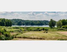 Weite, grüne Wiesen und Büsche im Vordergrund, dahinter ein glitzernder See mit Booten und dichter Waldlandschaft am Ufer; der Himmel ist teils sonnig mit einigen Wolken.
