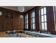 Ein historischer Raum mit Holzwänden, hohen Fenstern mit Sprossen und großen runden Hängelampen. Der Raum ist möbliert mit einfachen Holztischen und Plastikstühlen; an einer Wand ist ein Gemälde zu sehen.