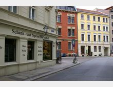 Straßenszene mit Altbauten, darunter ein beiges Gebäude mit der Aufschrift „Schank- und Speisewirtschaft“ und großen Fenstern. Im Hintergrund stehen ein rotes und ein gelbes Gebäude mit historischen Fassaden, während Fahrräder und ein gepflasterter Gehweg den Straßenrand säumen.