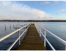 Ein langer, schmaler Holzsteg mit weißem Geländer führt über einen ruhigen See. Im Hintergrund sind eine bewaldete Uferlinie, vereinzelte Gebäude und ein teils bewölkter, blauer Himmel zu sehen.