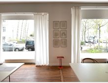 Ein heller Raum mit Holzboden, großen Fenstern mit weißen Vorhängen und zwei Tischen. An der weißen Wand hängen sechs gerahmte Zeichnungen und ein kleiner, roter Beistelltisch steht in der Mitte. Draußen sind parkende Autos, Bäume und Wohnhäuser zu sehen.